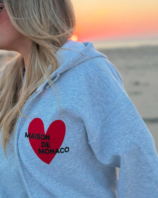 Person wearing a grey zip up hoodie with a red heart and 'MAISON DE MONACO' text on a beach at sunset.