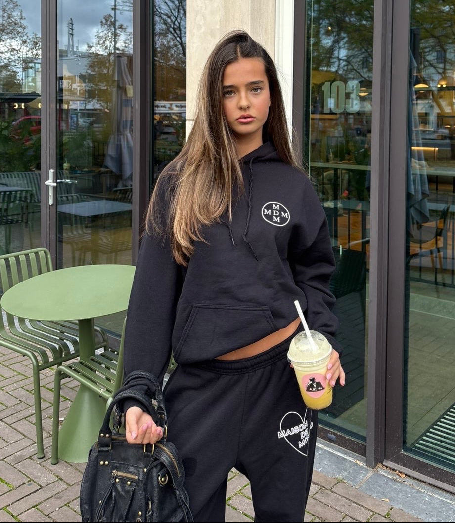 Woman in black hoodie and pants holding a drink outside a building with glass doors.