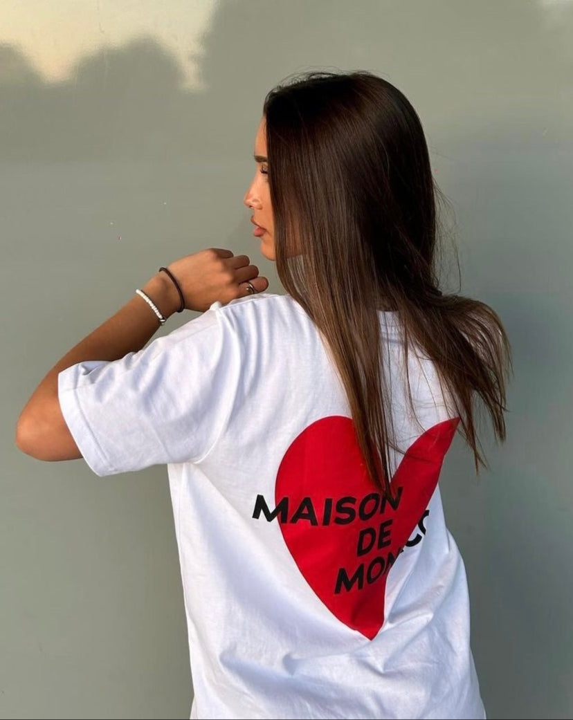 Person wearing a white t-shirt with a red heart and text, standing against a light gray wall.